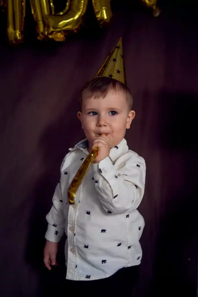 Jeune enfant avec un chapeau de fête doré soufflant dans un kazoo, devant une décoration d'anniversaire scintillante.