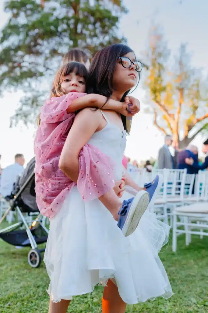 Deux jeunes filles en robe blanche, l'une portant l'autre sur son dos, profitant d'un mariage en extérieur avec des invités en arrière-plan.