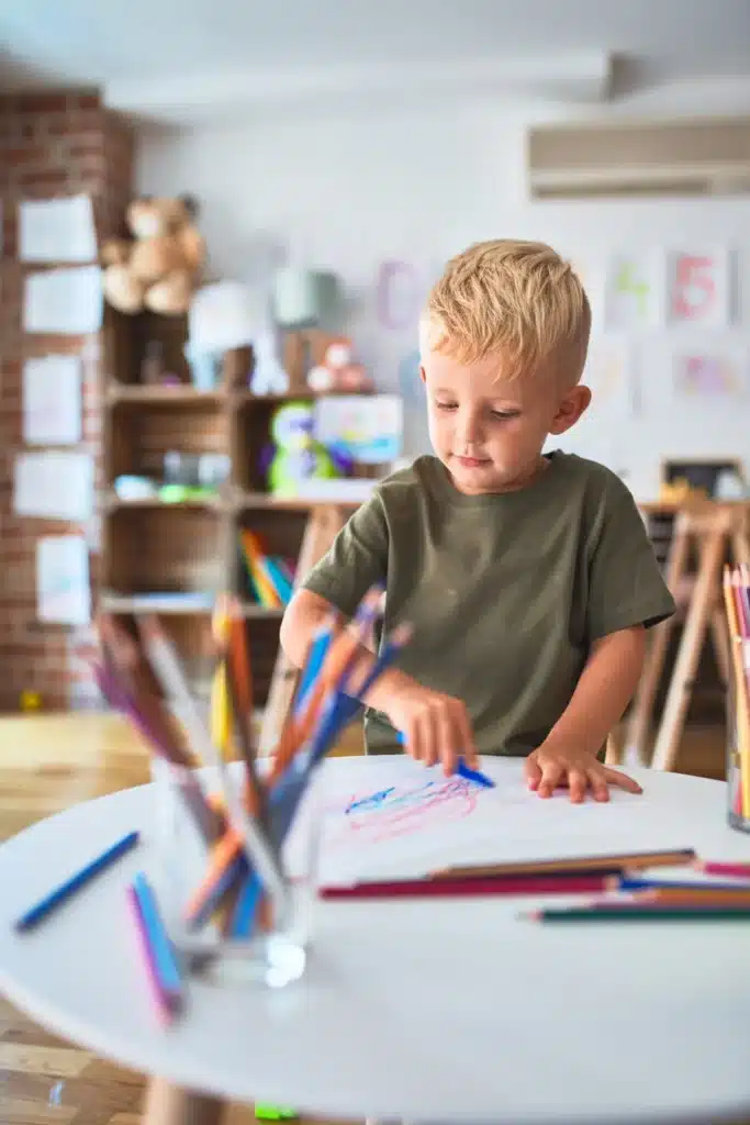 Un jeune garçon blond dessine avec des crayons de couleur sur une table blanche, dans un environnement d’apprentissage chaleureux et créatif.