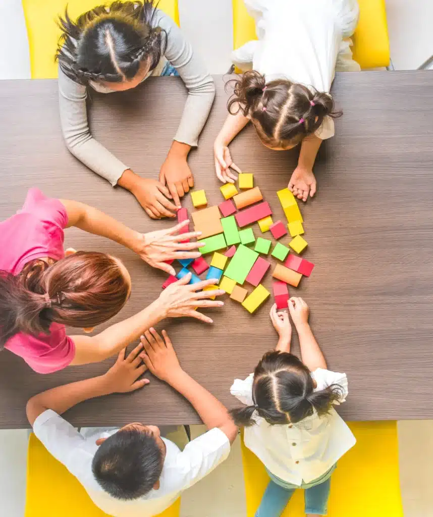 Vue de dessus d'une nourrice encadrant des enfants autour d'une table, en train de jouer avec des blocs colorés.